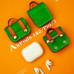 Può includere: Custodie AirPods in pelle verde con dettagli e manici marroni, insieme a una custodia e auricolari AirPods bianchi. Le custodie sono etichettate "AirPods 1&2/Pro/3" e presentano il testo "HANDCRAFTED LEATHER BY NEEDLEWORK". Lo sfondo è arancione.