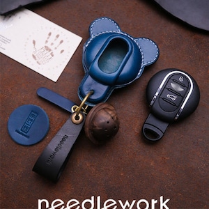 Può includere: Una custodia per portachiavi in pelle blu a forma di orso, un portachiavi in pelle nera e una perlina di legno. L'immagine include anche una chiave dell'auto e un piccolo cerchio di pelle blu. La parola "needlework" è stampata sul portachiavi.