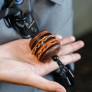 Puede incluir: Un llavero de cuero artesanal con un diseño único. El llavero presenta un diseño circular apilado con capas alternas de cuero marrón oscuro, negro y marrón claro. Incluye un mosquetón negro y un cordón negro con dos extremos cilíndricos.