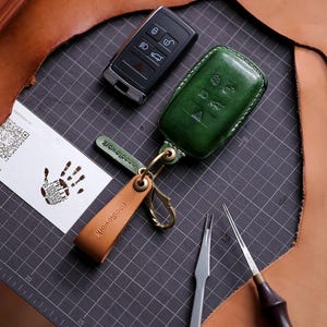 May include: A green leather key fob cover and a tan leather keychain with the word "needlework" embossed. A black car key and a business card with a handprint are also present. The items are on a grey cutting mat.