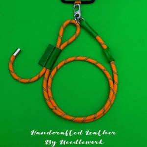 Może przedstawiać: Pomarańczowa i szara smycz do telefonu z liną z zielonymi skórzanymi akcentami i srebrnym zapięciem. Smycz jest przymocowana do czarnego etui na telefon. Tekst "Handcrafted Leather By Needlework" znajduje się na dole.