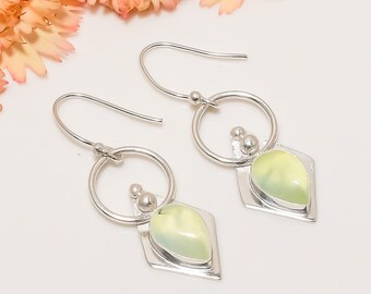 May include: A pair of silver earrings with a light green, teardrop-shaped stone set in a silver frame. The earrings have a circular design with small silver beads and a hook closure. The background is white with a hint of pink flowers.