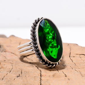 Puede incluir: Un anillo de plata con una gran piedra preciosa verde brillante de corte ovalado. La piedra preciosa está engastada en un bisel texturizado y detallado. La banda del anillo tiene un diseño de triple barra. El anillo se muestra sobre una superficie de madera.