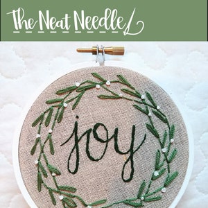 May include: A round, beige fabric embroidery hoop with the word "joy" stitched in black script. The word is surrounded by a green embroidered wreath with white bead accents. The text "Joy Christmas Ornament" is below the hoop.