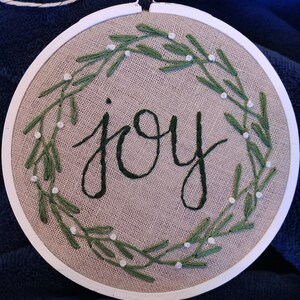 May include: Embroidered hoop art with the word "joy" in dark green script. The word is surrounded by a wreath of green leaves and small white beads, all stitched on a tan fabric. The hoop is white and has a gold-colored screw.
