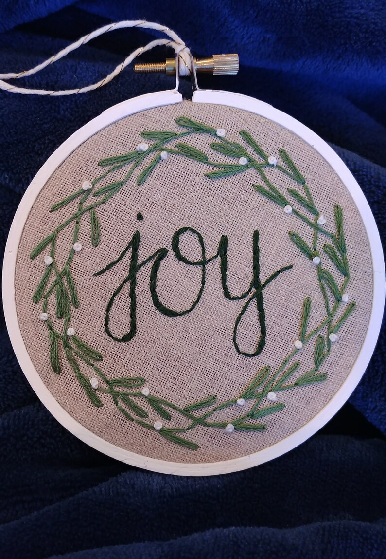 May include: Embroidered hoop art with the word "joy" in dark green script. The word is surrounded by a wreath of green leaves and small white beads, all stitched on a tan fabric. The hoop is white and has a gold-colored screw.
