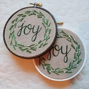 May include: Two round embroidery hoops with the word "joy" stitched in green cursive. Each hoop features a green embroidered wreath with white accents. One hoop has a dark brown frame, the other a white frame.