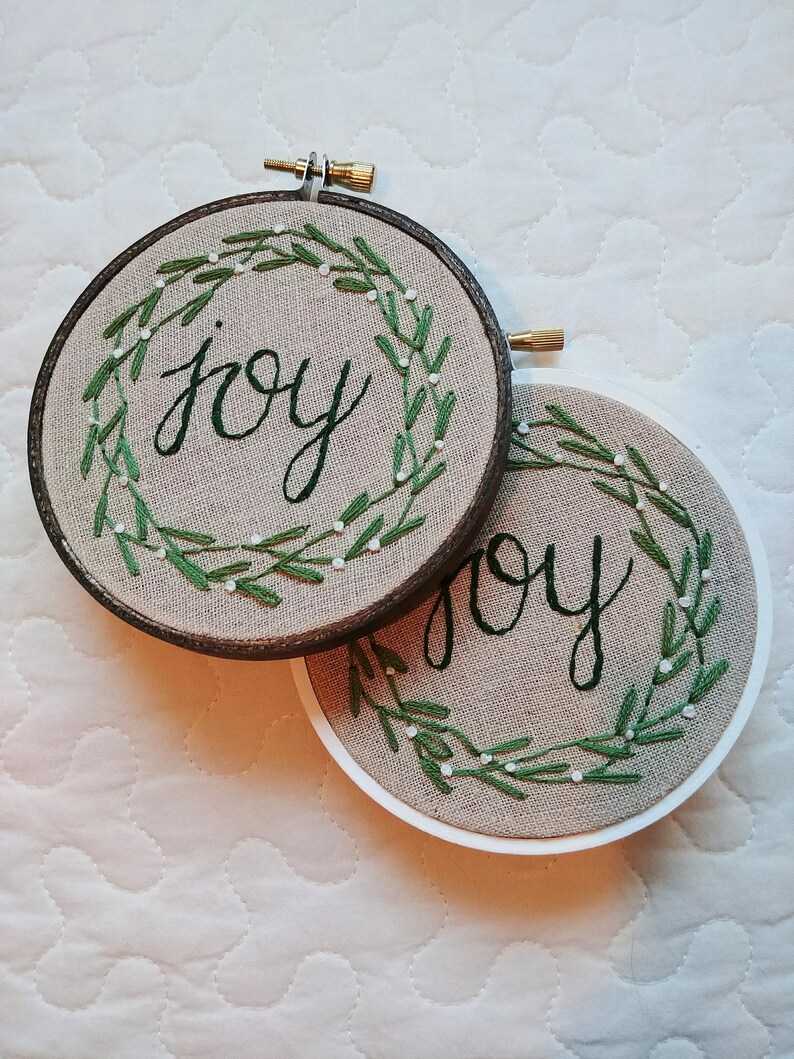 May include: Two round embroidery hoops with the word "joy" stitched in green cursive. Each hoop features a green embroidered wreath with white accents. One hoop has a dark brown frame, the other a white frame.