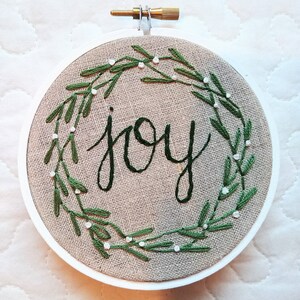 May include: Embroidered hoop art with the word "joy" in dark green script. The word is surrounded by a green leaf wreath with small white accents, all on a tan fabric background. The hoop is white with a brass screw.