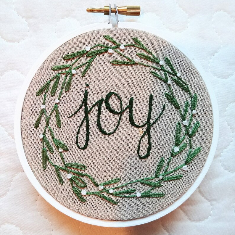 May include: Embroidered hoop art with the word "joy" in dark green script. The word is surrounded by a green leaf wreath with small white accents, all on a tan fabric background. The hoop is white with a brass screw.
