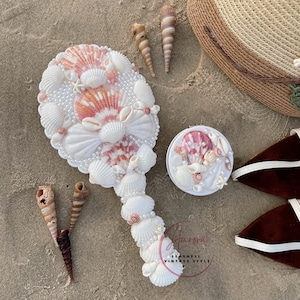 May include: A decorative hand mirror and compact mirror, both decorated with seashells and faux pearls. The mirror has a white handle and a shell-covered face with pink and white shells. A straw hat and seashells are also visible.