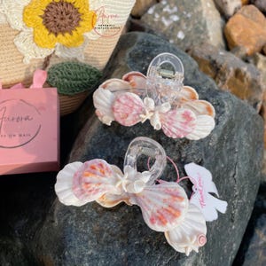 Könnte beinhalten: Zwei Muschel-Haarspangen mit transparenten Kunststoffverschlüssen. Die Spangen zeigen weiße und rosa Muscheln, die fächerförmig angeordnet sind. Ein kleines weißes Etikett mit rosa Text "Thank you!" ist befestigt.