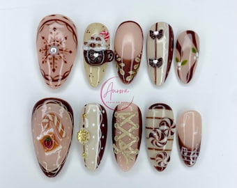 May include: A set of ten almond-shaped false nails with various designs. The nails feature a color palette of brown, beige, and white, with accents like snowflakes, bows, and musical notes. The word "Aurora" is written on the image.