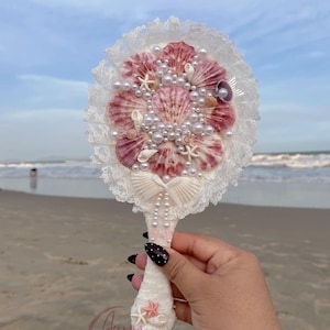 May include: A seashell-adorned hand mirror with a vintage style. The mirror features a cluster of pink and white seashells, pearls, and starfish, framed by white lace. The handle is decorated with seashells and a bow. The text "Aurora Seashell Vintage Style" is visible.