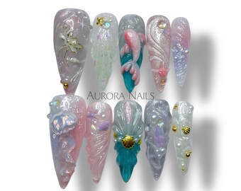 May include: A set of ten long, pointed acrylic nails with a mermaid theme. The nails feature various designs, including sculpted flowers, seashells, and aquatic creatures, in shades of pink, teal, and iridescent white. The text "AURORA NAILS" is visible.