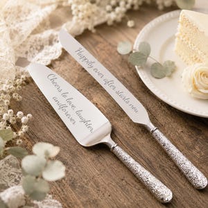 May include: Silver cake knife and server set with engraved text. The knife reads "Happily ever after starts now," and the server reads "Cheers to love, laughter, and forever." The handles have a textured floral design.