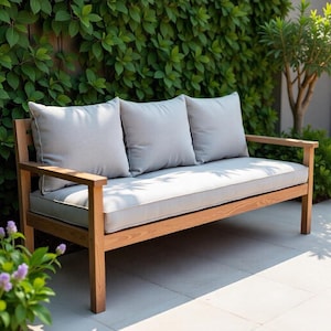 Può includere: Una panca da giardino in legno con tre cuscini e cuscini grigi. La panca ha una struttura marrone chiaro. I cuscini e i cuscini sono di colore grigio chiaro con bordatura bianca. La panca è ambientata su uno sfondo di vegetazione verde e un alberello.
