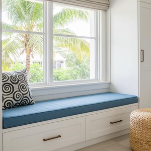 Könnte beinhalten: Fenstersitz mit blauem Kissen und weißen Schubladen. Ein dekoratives Kissen mit einem schwarz-weißen Wirbelmuster liegt auf dem Sitz. Ein gewebter Hocker steht daneben. Das Fenster blickt auf eine tropische Landschaft.