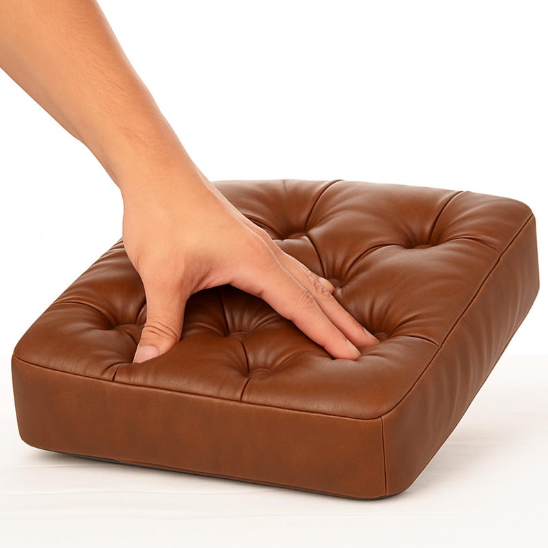Op de afbeelding: Een bruin, vierkant kussen met een gecapitonneerd ontwerp. Het kussen is gemaakt van een glad materiaal en heeft een zachte uitstraling. Een hand drukt op het kussen.
