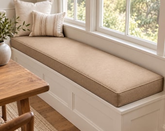 May include: A long, rectangular bench cushion in a solid tan fabric with piped edges, placed atop a white built-in window seat. Two decorative throw pillows, one solid tan and one tan with vertical stripes, rest against the window frame.
