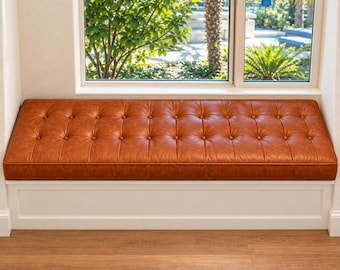 May include: A long rectangular window bench featuring a tufted cognac-colored leather cushion with button detailing. The bench is supported by a solid white base with clean lines, positioned directly beneath a large window with a view of greenery.