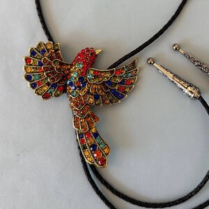 May include: A bolo tie featuring a colorful jeweled bird with outstretched wings. The bird is adorned with red, blue, yellow, and orange gemstones. The tie has a black braided cord and silver-toned tips.