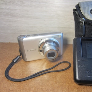May include: A silver Canon digital camera with a retractable lens, a black wrist strap, and a black charger. A gray and black Lowepro camera case is also included.