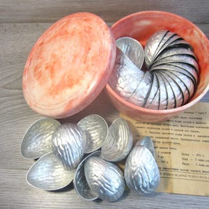May include: A vintage, pink and white container with a lid, filled with silver-colored, egg-shaped molds. Several molds are also outside the container. A piece of paper with text is visible.