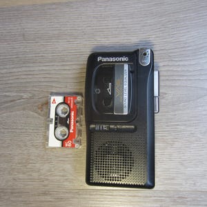 May include: A black Panasonic microcassette recorder with a built-in speaker, a microphone port, and a VAS sound level equalizer feature, positioned next to a small Panasonic MC-60 microcassette tape with a red and white label.