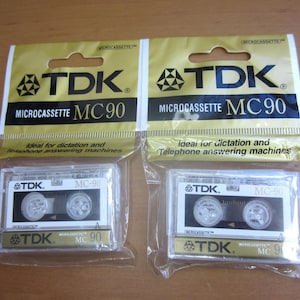 May include: Two TDK Microcassette MC90 tapes in their original packaging. The packaging is gold and black with the TDK logo and product information. The clear plastic cases show the tape reels.