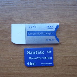 Op de afbeelding: Een witte en zilveren Sony Memory Stick Duo Adapter naast een blauwe SanDisk Memory Stick Pro Duo 1GB kaart. De adapter heeft de tekst "Memory Stick Duo Adaptor" en de kaart "MEMORY STICK PRO DUO".