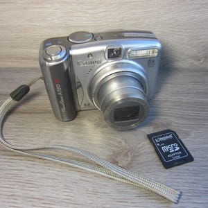 May include: Silver Canon PowerShot A720 IS digital camera with a 6x zoom lens and a black Kingston Micro SD card. The camera has a silver and black wrist strap.