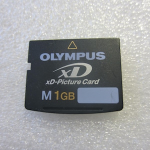 Puede incluir: Tarjeta xD-Picture Olympus negra con texto blanco y dorado. La tarjeta tiene el logotipo de Olympus, el texto "xD-Picture Card" y "M 1GB". La tarjeta es rectangular con un borde dentado.