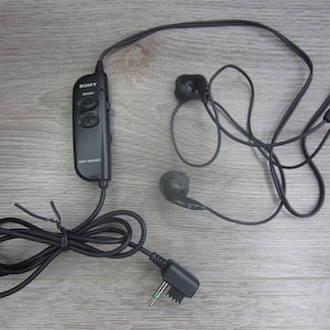 May include: Black in-ear headphones with a control module. The headphones have a 3.5mm jack and are labelled "SONY". The control module has "REPEAT" and two other buttons. The headphones are on a wooden surface.