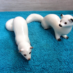 Puede incluir: Dos figuras de cerámica blanca de comadrejas. Una está tumbada, la otra de pie. Ambas tienen detalles negros en las orejas, los pies y los ojos. Las figuras están sobre una superficie azul texturizada.