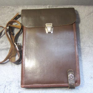 May include: A brown leather satchel with a flap closure and a shoulder strap. The satchel has a rectangular shape with a silver metal clasp and a smaller strap closure on the bottom. The shoulder strap is adjustable.