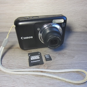 May include: A black Canon digital camera with a silver lens and flash. The camera has a wrist strap and is accompanied by a black SD card adapter and a smaller SD card. The camera lens reads "Canon Zoom Lens 3.3x".