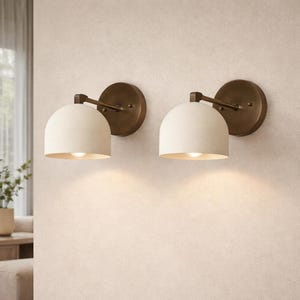 May include: Two wall sconces with a cream-colored, dome-shaped shade and a warm, bronze-toned metal base. The sconces are mounted on a light-colored wall, casting a soft glow.
