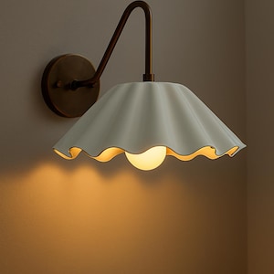 May include: A wall-mounted light fixture with a white, scalloped shade and a warm, glowing bulb. The fixture has a bronze-colored arm and a circular base, creating a vintage aesthetic. The light casts a soft, inviting glow on the wall.