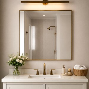 May include: A bathroom vanity featuring a white countertop, a rectangular sink, and a brass faucet. Above the sink is a square mirror with a brass frame and a horizontal brass light fixture. A vase of white roses and a woven basket are present.