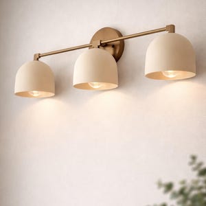 May include: A three-light vanity fixture with a brass finish. The light shades are a cream color and dome-shaped. The fixture is mounted on a wall and has a warm glow from the light bulbs.