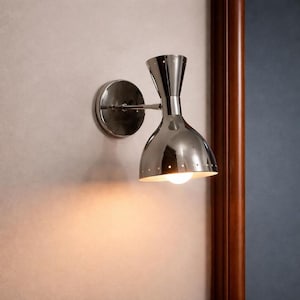 Puede incluir: Aplique de pared cromado pulido con una pantalla cónica y una placa de montaje redonda. La lámpara tiene un diseño clásico y moderno de mediados de siglo, y está montada en una pared de color claro. El aplique está iluminado, proyectando un brillo cálido.