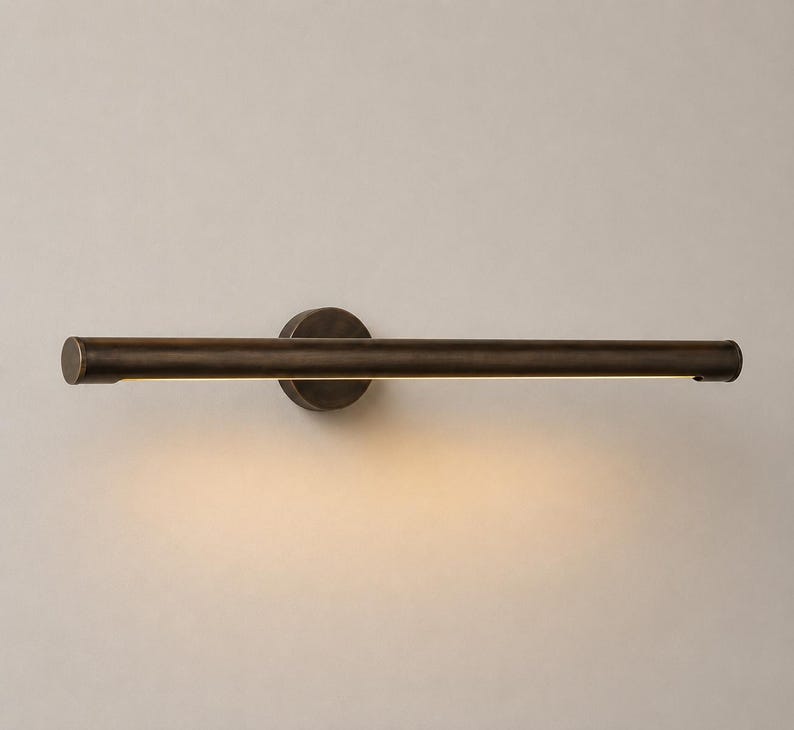 May include: A horizontal wall-mounted light fixture with a dark bronze finish. The cylindrical bar features a central circular mounting plate and emits a warm, soft glow from the underside, illuminating the space directly below the fixture.