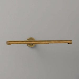 May include: A horizontal, tubular picture light fixture finished in an antique brass tone. The light features a central circular wall-mounting plate that connects to a long, slender cylindrical bar with capped ends, designed for illuminating wall art.