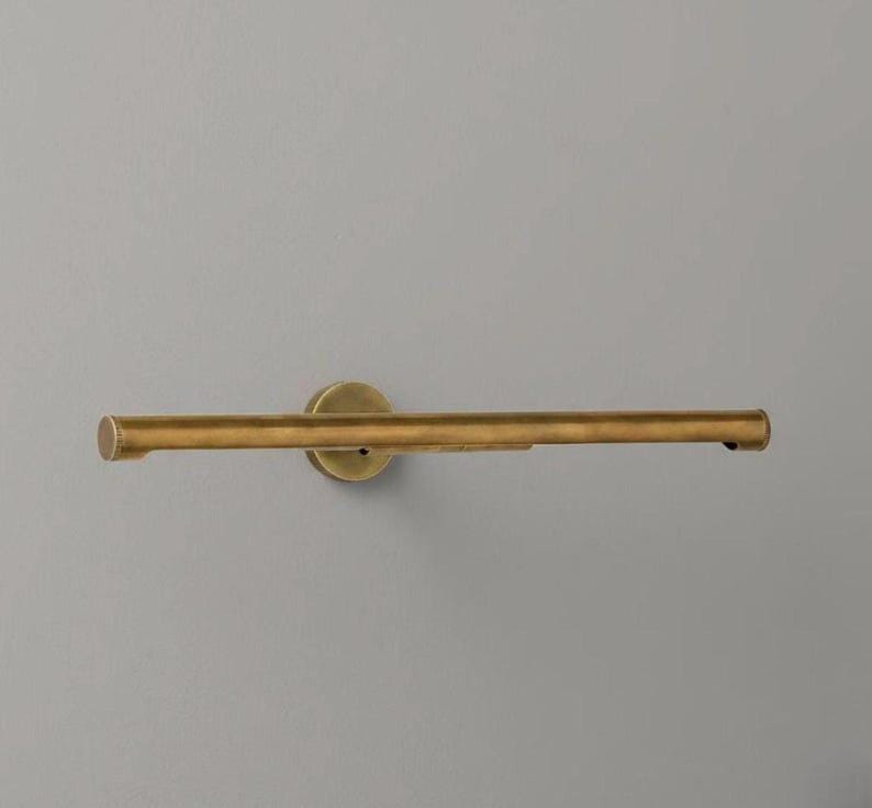 May include: A horizontal, tubular picture light fixture finished in an antique brass tone. The light features a central circular wall-mounting plate that connects to a long, slender cylindrical bar with capped ends, designed for illuminating wall art.