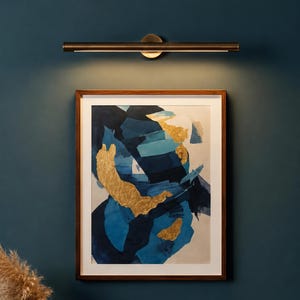 May include: A rectangular framed wall art piece featuring abstract brushstrokes in shades of navy blue, light blue, and white, accented with gold leaf patches. Above the artwork is a horizontal, cylindrical metal wall light fixture.