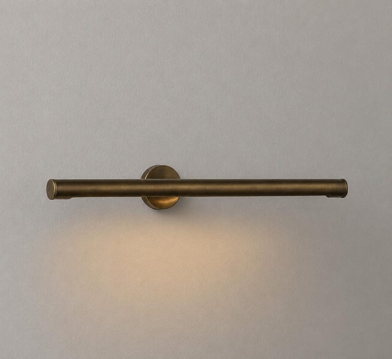May include: A horizontal wall-mounted light fixture with a cylindrical, antique brass-toned metal bar. The fixture features a circular base plate that attaches to the wall, and it emits a soft, warm downward glow from the underside of the tube.