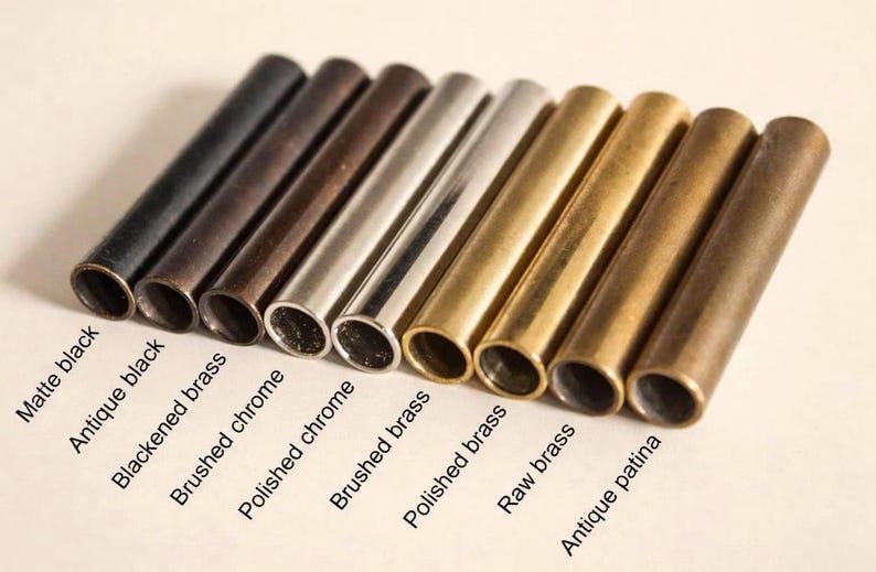 May include: A row of nine cylindrical metal tubes in various finishes, labeled as Matte black, Antique black, Blackened brass, Brushed chrome, Polished chrome, Brushed brass, Polished brass, Raw brass, and Antique patina, arranged side by side.