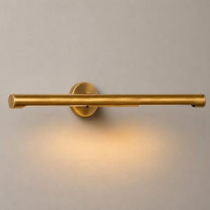 May include: A horizontal wall-mounted picture light with a cylindrical brass finish tube. The fixture features a circular base plate and emits a warm, soft glow directed downward from the underside of the metal bar, illuminating the area below.