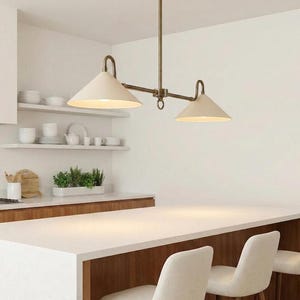 May include: A two-light pendant light fitting with a brass finish and off-white cone-shaped shades. The fitting is suspended from the ceiling, illuminating a kitchen island. The kitchen has white countertops and wooden cabinets.